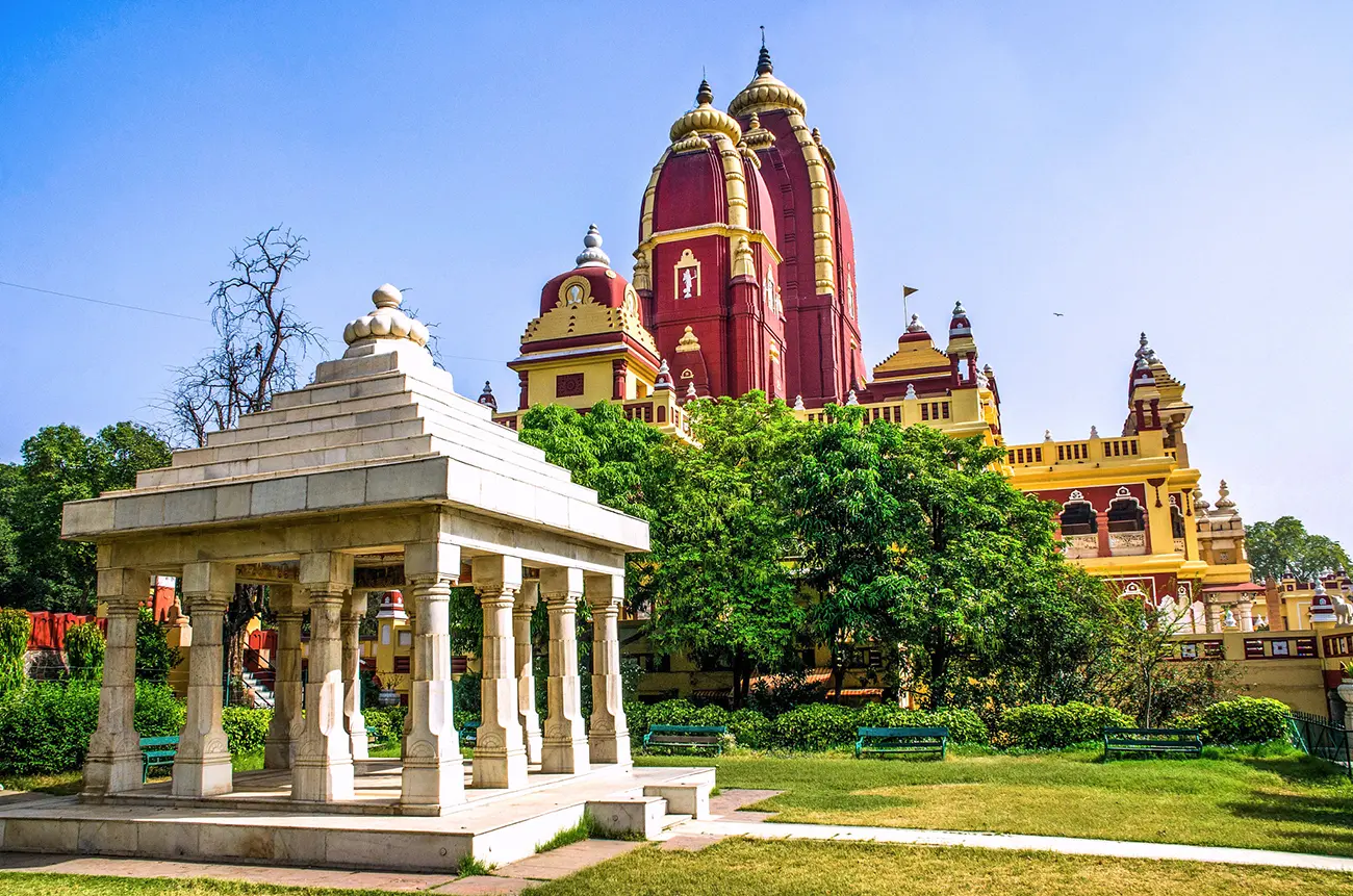 The vibrant Birla Temple in India, with its prominent red and cream spires and domes, stands beside an urban road with a fence, under a clear blue sky.