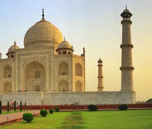 Taj Mahal at sunset, a white marble mausoleum with four minarets, surrounded by green lawns and red sandstone buildings under a golden sky.