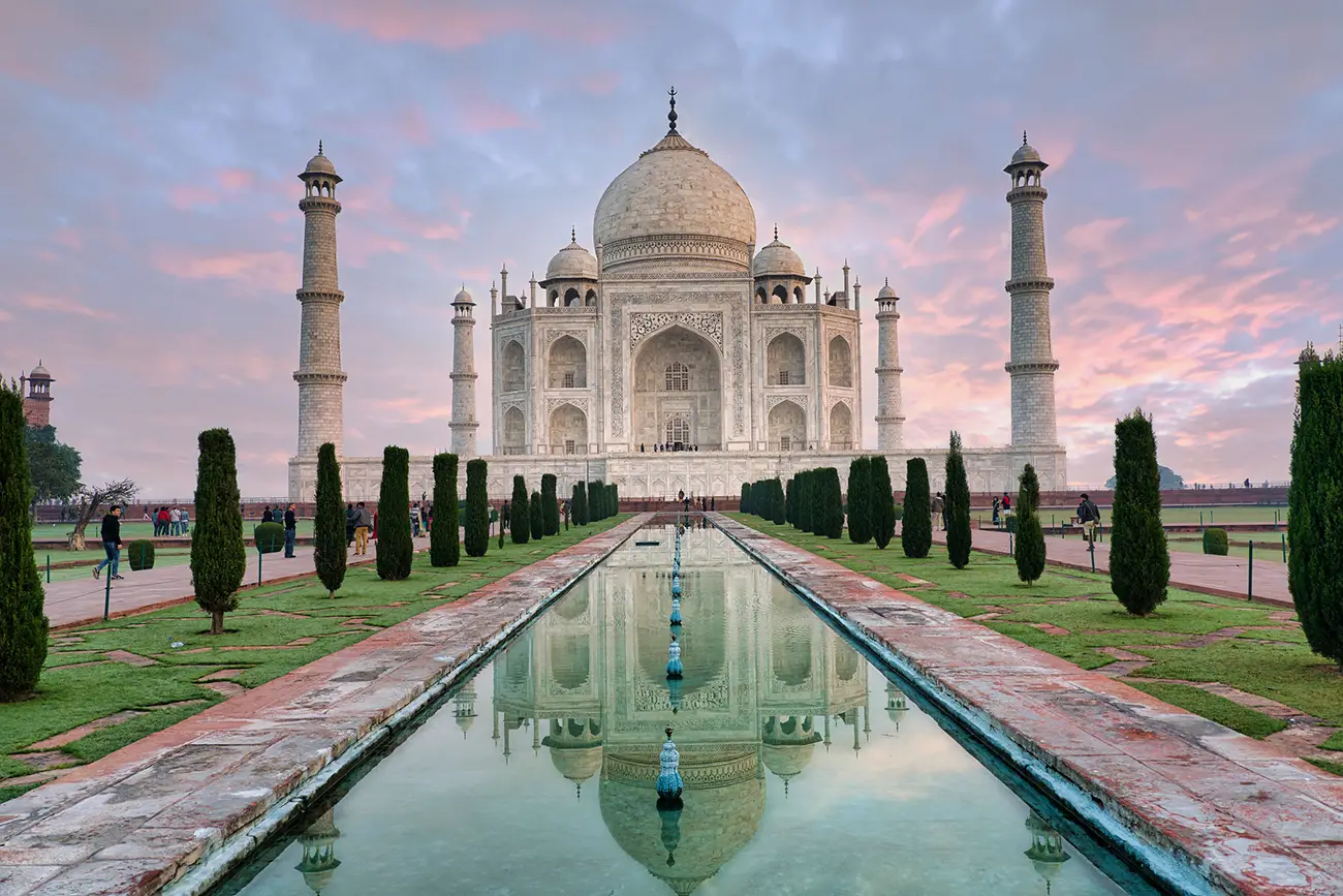 The Taj Mahal reflected in its long rectangular pool, with the white marble mausoleum and its minarets under a soft, cloudy sky.
