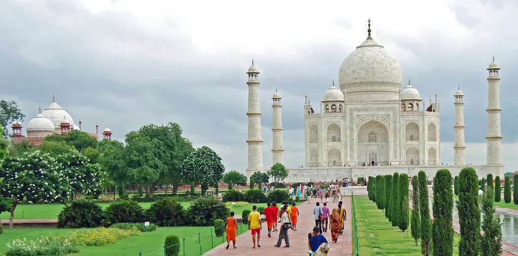 The Taj Mahal, a grand white marble mausoleum, seen from a distance with visitors walking along a pathway lined with green hedges and trees, under a cloudy sky.