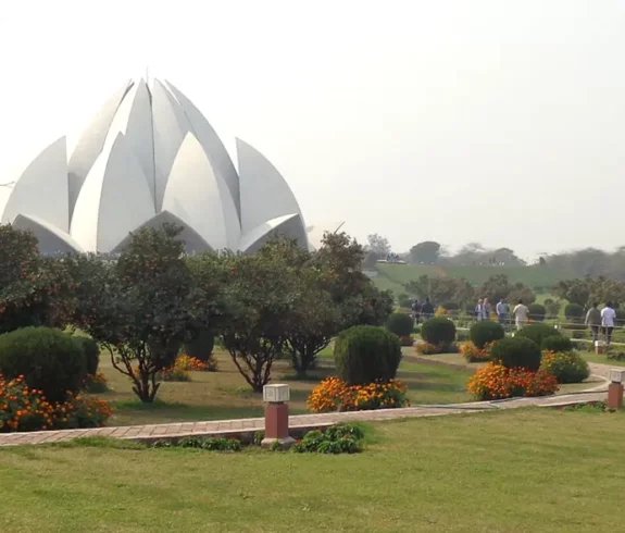 A panoramic view of the white, lotus-shaped Baháʼí House of Worship (Lotus Temple) in New Delhi, India, surrounded by lush green gardens, pathways, and trees under a clear sky.