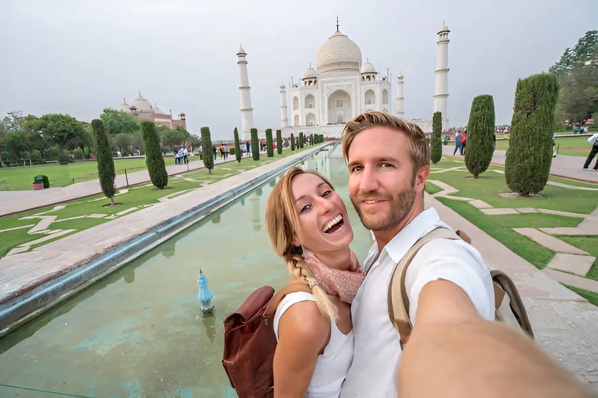 A happy couple takes a selfie in front of the Taj Mahal, which is visible in the background with its reflection pool and green lawns.