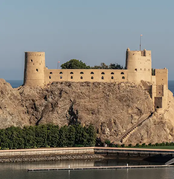 Al Jalali Fort in Muscat, Oman, stretching across a rocky cliff above the Arabian Sea with round watchtowers and sand-colored walls.