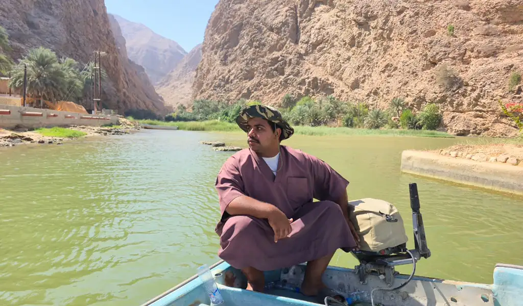 A man sitting on a small boat during a scenic boat ride in Wadi Shab, Oman, surrounded by rocky canyon cliffs and green water.