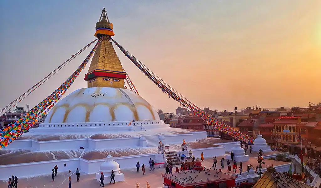 Boudhanath Stupa in Kathmandu, Nepal, with a large white dome, golden spire, Buddha eyes, and colorful prayer flags at sunset.