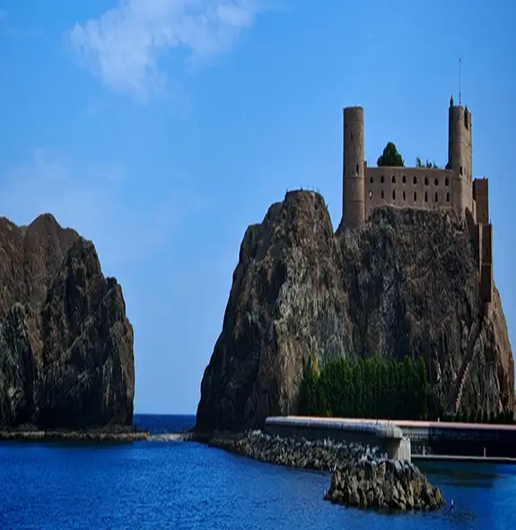 A stone fortress sits atop a rocky coastal headland beside calm blue water under a clear sky.