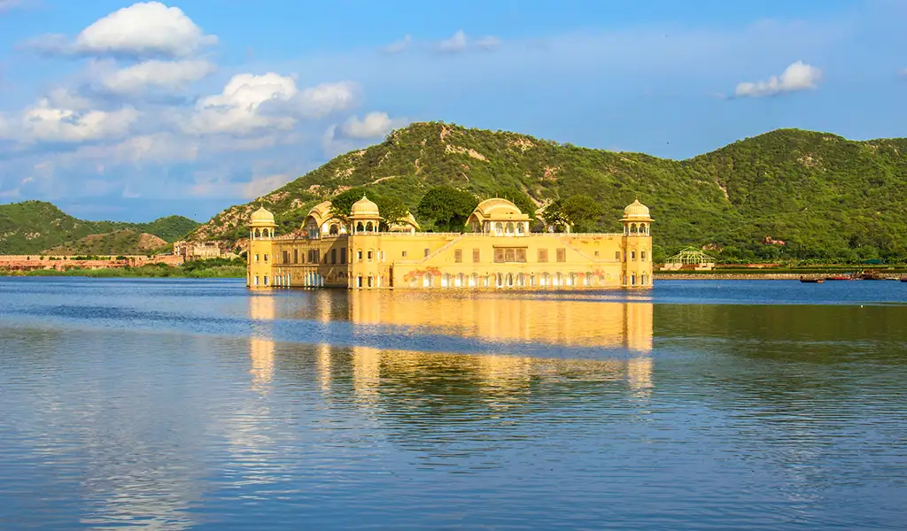 Jal Mahal, a yellow sandstone palace, sits in the middle of Man Sagar Lake in Jaipur, India, with green hills and a blue sky behind.