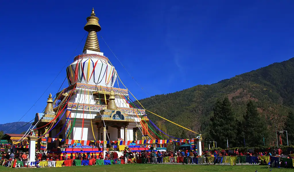 A large white Buddhist stupa decorated with colorful prayer flags, with a crowd gathered in front and forested mountains under a deep blue sky.