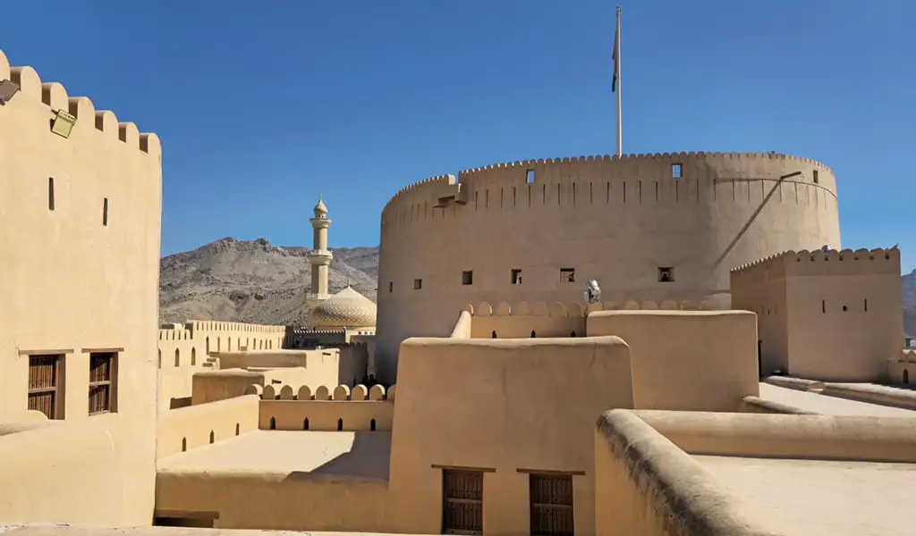 Nizwa Fort in Oman with its large round tower, traditional mud-brick walls, and mountain backdrop under a clear blue sky.