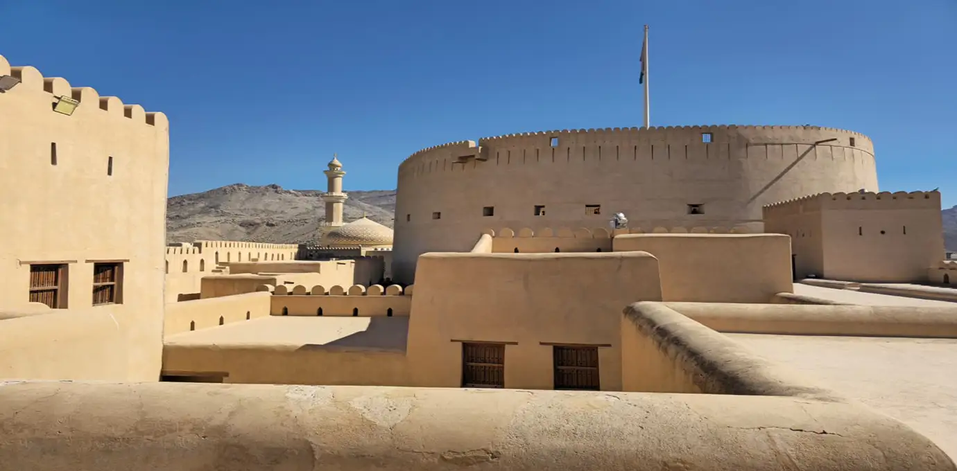 Nizwa Fort in Oman during Oman Highlight Tour with its large round tower, adobe walls, and mountain backdrop under a clear blue sky.