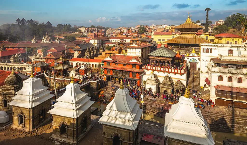 Pashupatinath Temple complex in Kathmandu, Nepal, with white shrines in the foreground and a crowded riverside temple area filled with colorful buildings.