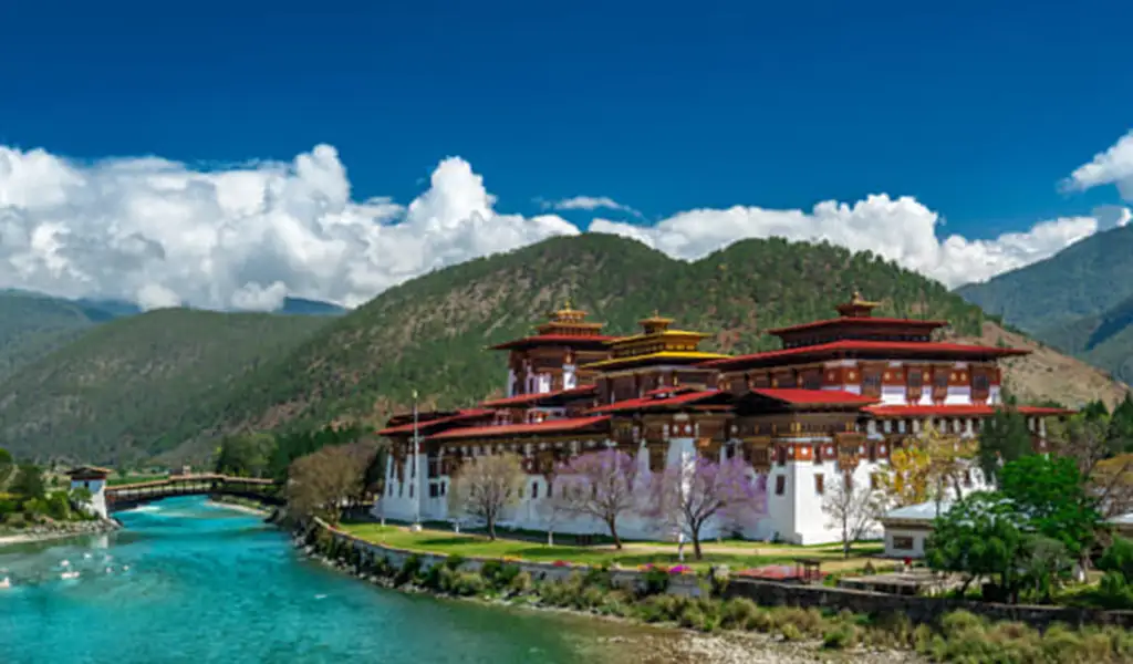 Punakha Dzong, a large white fortress with red and gold roofs, sits beside a turquoise river with green mountains and clouds behind.
