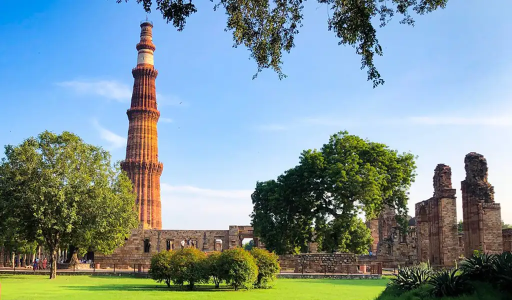 Qutub Minar rises above green lawns and trees in Delhi, India, with historic stone ruins in the background under a clear blue sky.