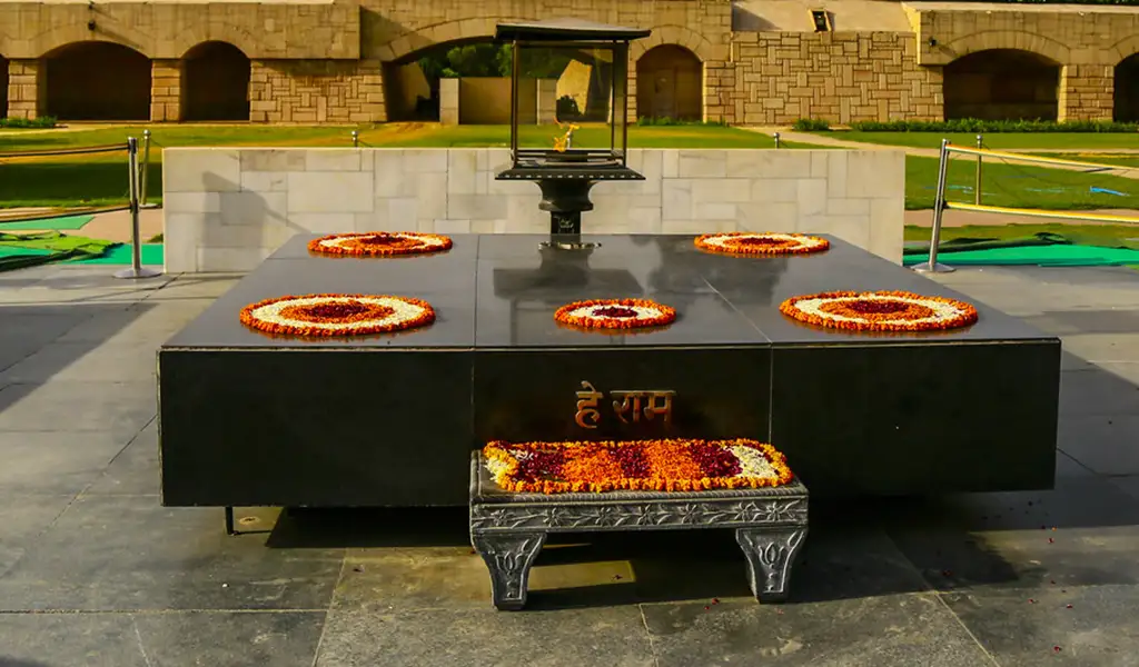A black stone memorial platform at Raj Ghat, decorated with orange and white flower wreaths, with an eternal flame burning at the center.