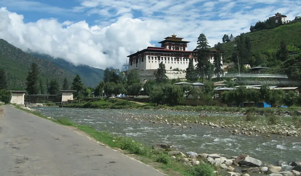 Rinpung Dzong fortress-monastery stands on a hillside above a river valley in Paro, Bhutan, with mountains and clouds in the background.