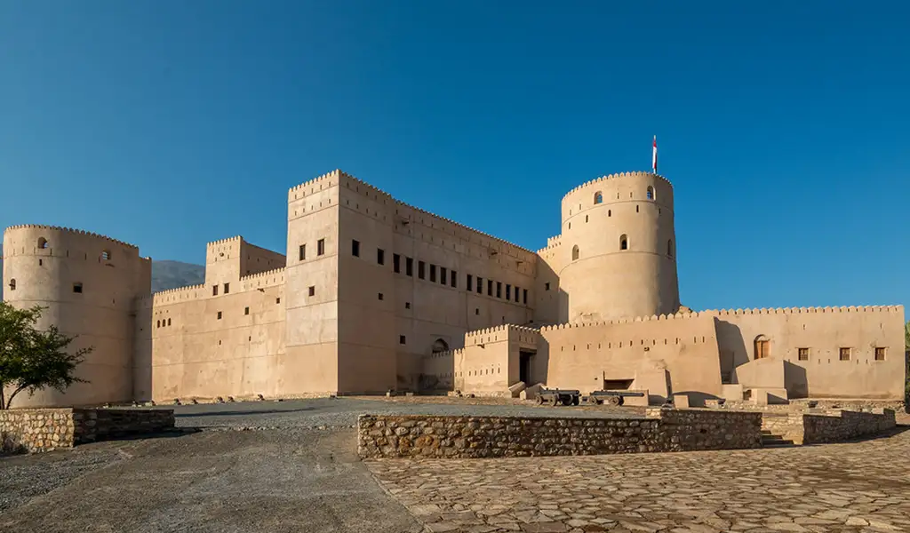 Rustaq Fort in Oman with massive sand-colored walls and a round tower, under a clear blue sky.