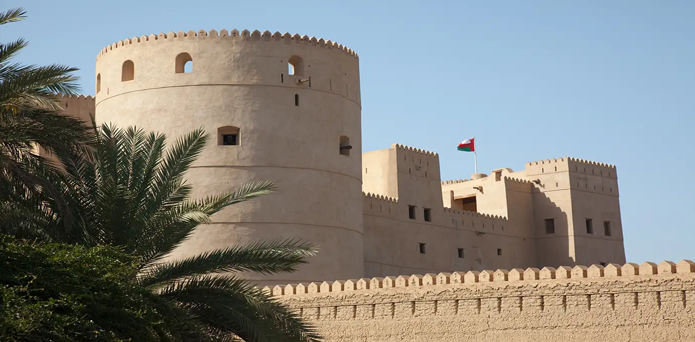 Rustaq Fort in Oman with palm trees in the foreground and the Omani flag on the tower.