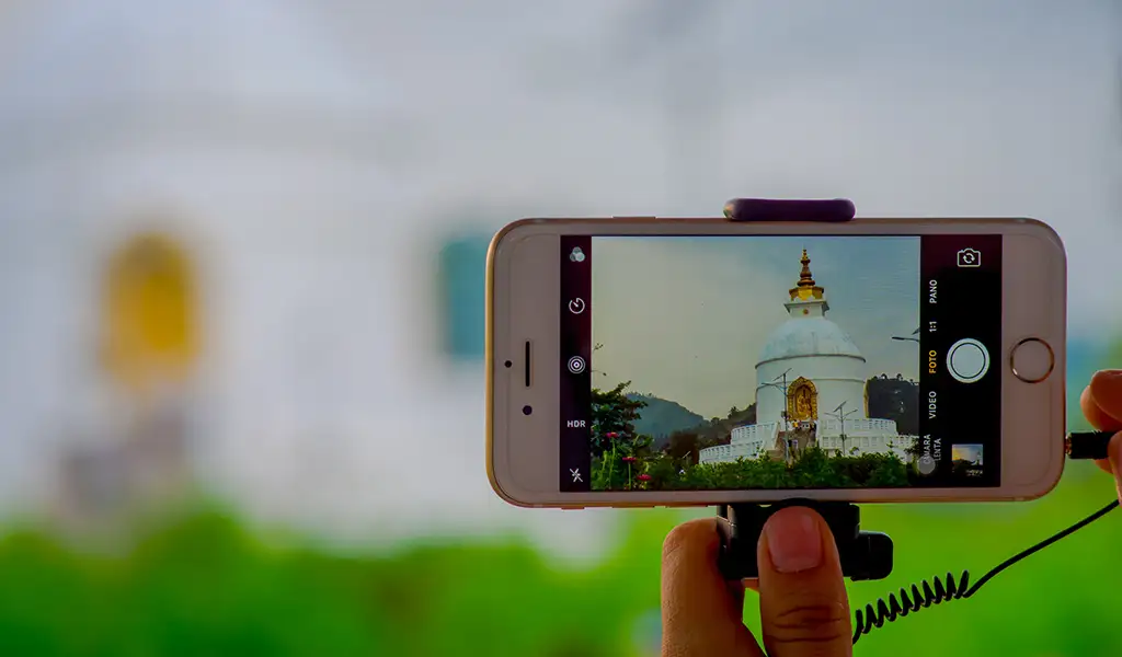 A person holds a smartphone taking a photo of the white Shanti Stupa in Pokhara, Nepal, with the stupa visible on the phone screen.