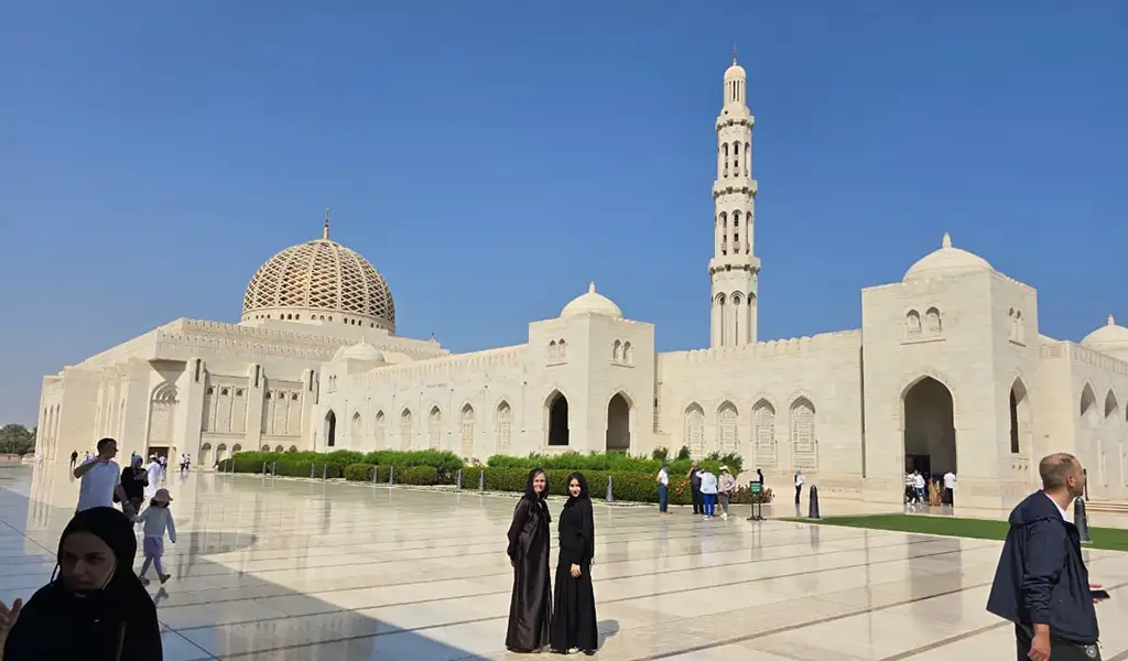 Sultan Qaboos Grand Mosque in Muscat, Oman, with its white marble courtyard, dome, and tall minaret under a clear blue sky.