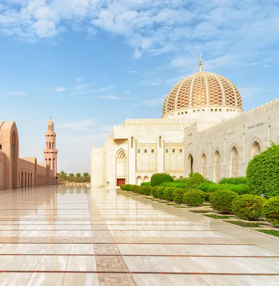 Courtyard view of the Sultan Qaboos Grand Mosque in Muscat, Oman, showing arched corridors, a white marble façade, and a large patterned dome under a clear sky.
