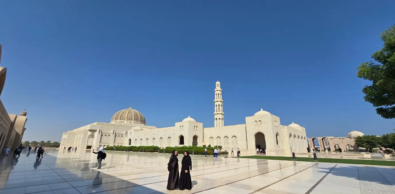 Sultan Qaboos Grand Mosque in Oman Highlights Tour with white marble architecture, domes, minaret, and visitors under a bright blue sky.