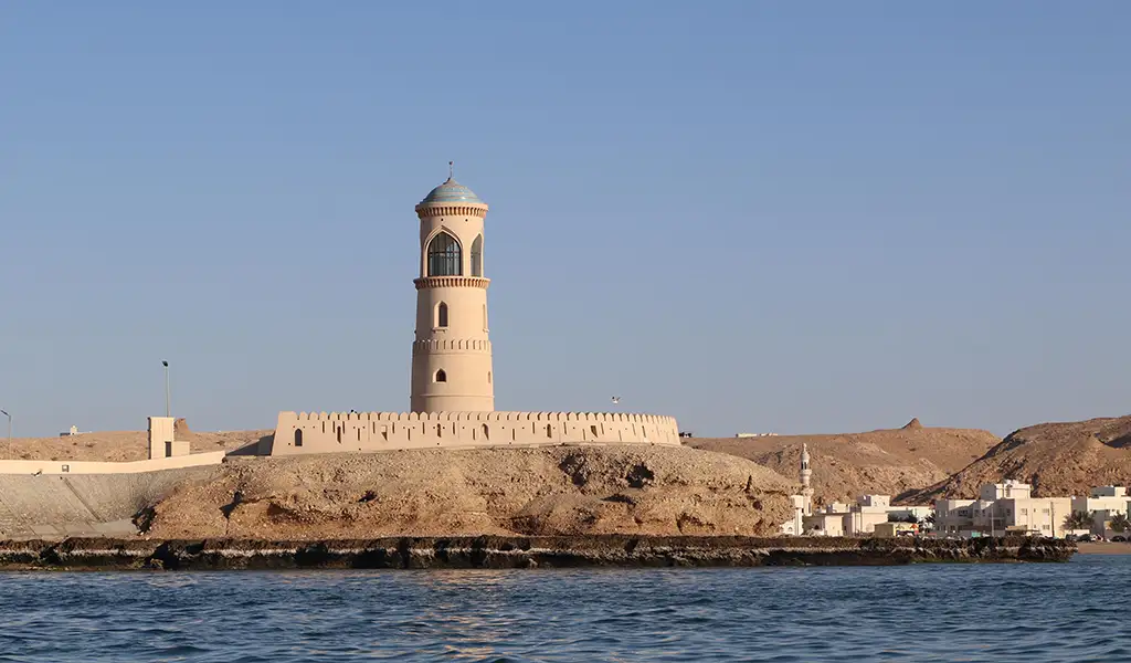 A tall beige lighthouse stands on a rocky shoreline in Sur, Oman, with blue sea in the foreground and desert hills behind.