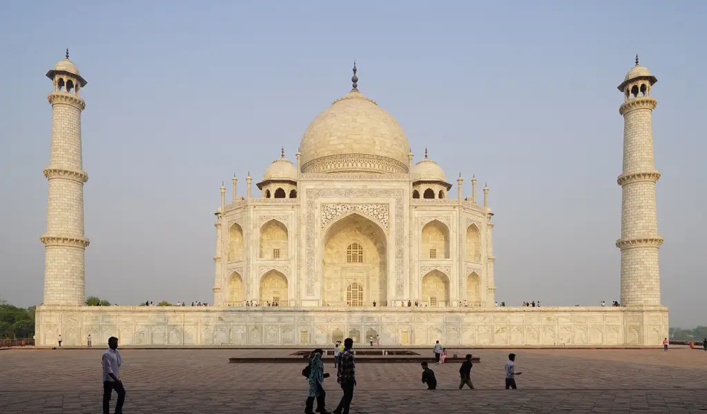 The Taj Mahal in Agra, India, with its white marble dome and minarets, viewed from the courtyard with visitors walking in the foreground.