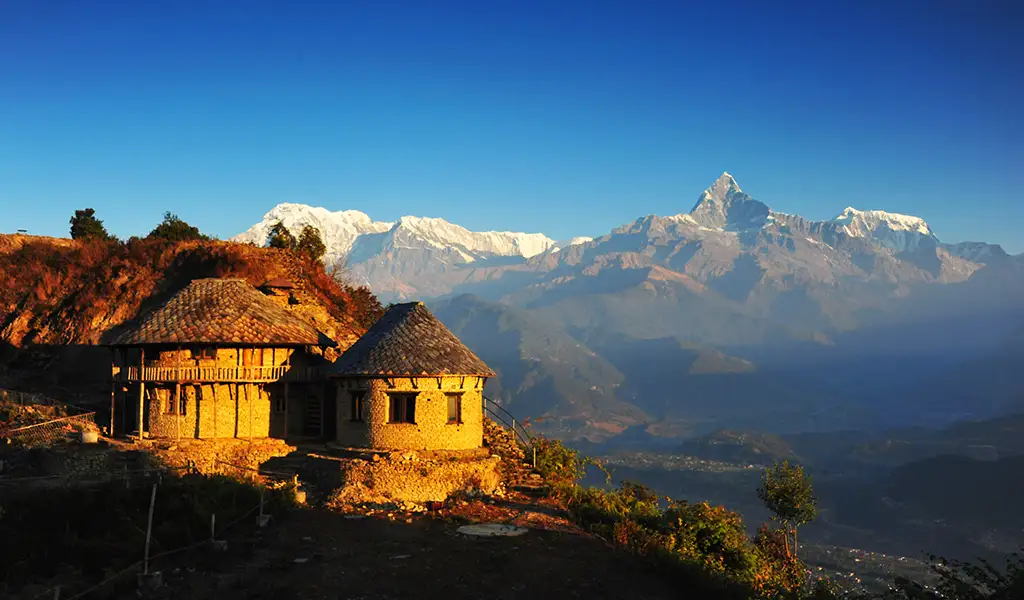 Stone village houses on a hillside overlook the Himalayas near Pokhara, Nepal, with snow-capped peaks under a clear blue sky.