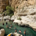 A diverse group of visitors swimming and wading in the clear, light-blue pool of Wadi Bani Khalid, surrounded by rugged, sun-bleached desert canyon walls in Oman.