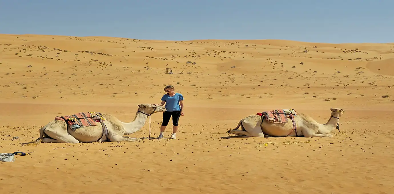 Oman Highlight Tour Woman standing beside two resting camels in the golden dunes of Wahiba Sands, Oman.