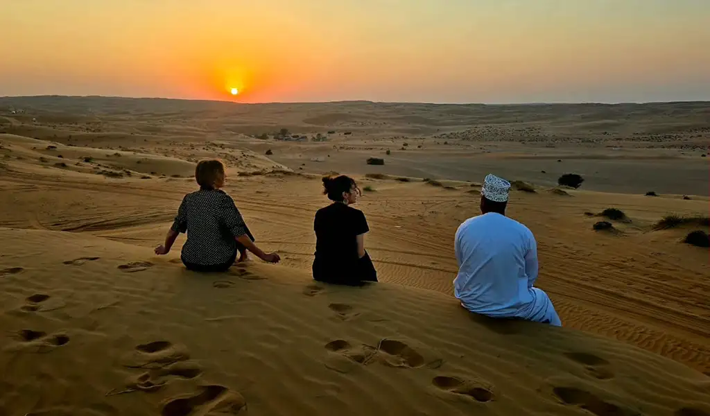 Travelers sitting on sand dunes watching the sunset in Wahiba Sands desert, Oman, with golden dunes stretching into the distance.