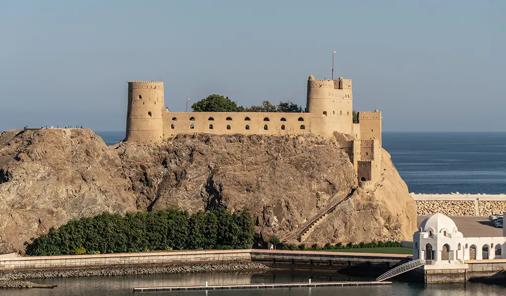 Al Jalali Fort in Muscat, Oman, stretching across a rocky cliff above the Arabian Sea with round watchtowers and sand-colored walls.