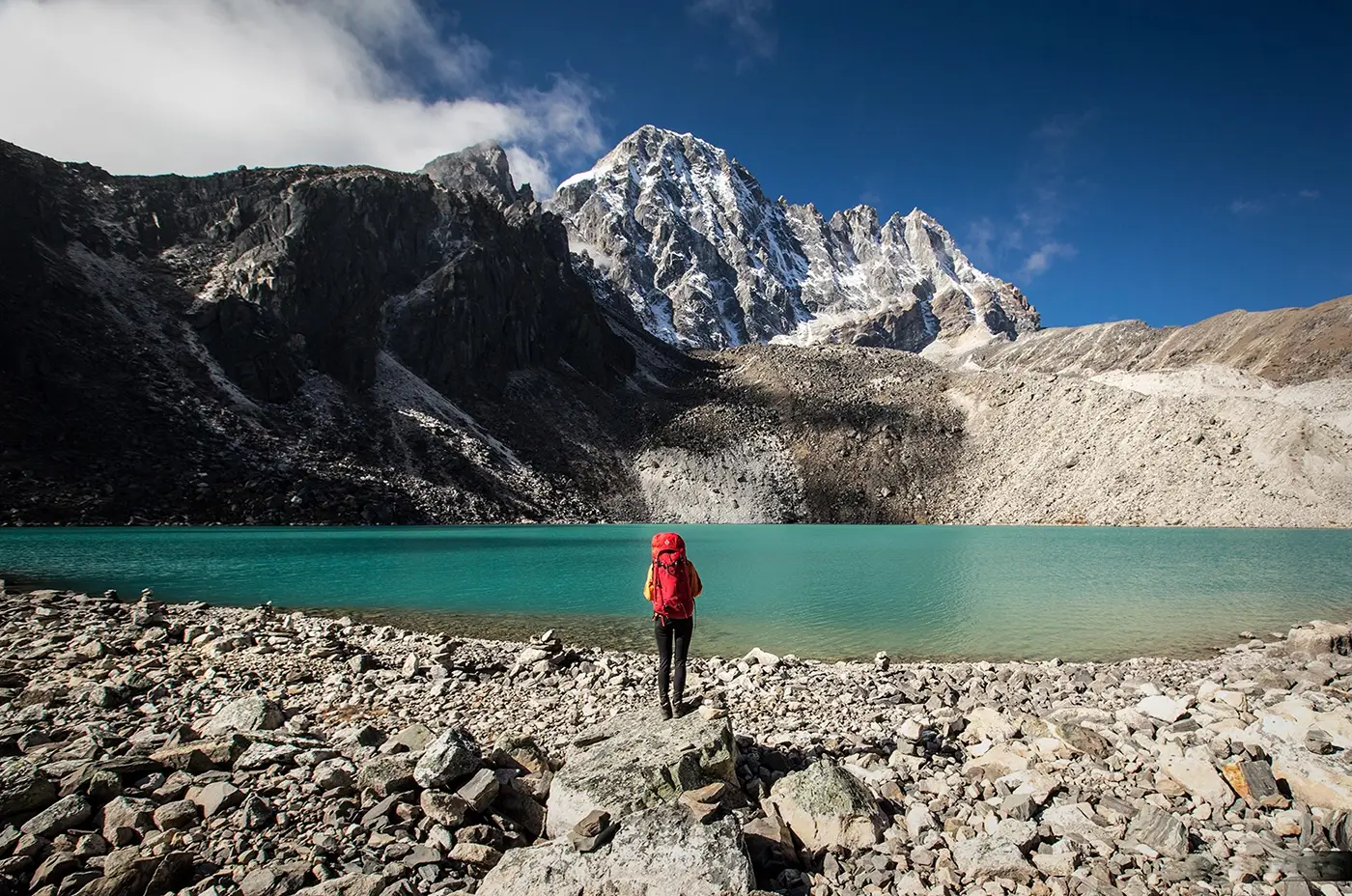 Trekker standing beside turquoise Gokyo Lake with snow-covered Himalayan peaks and rocky alpine landscape in Nepal