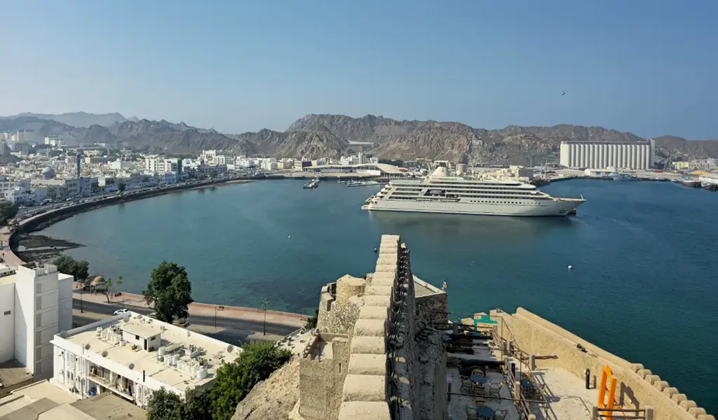 View of Mutrah Corniche in Muscat, Oman, with a cruise ship in the harbor, waterfront buildings, and mountains in the background.