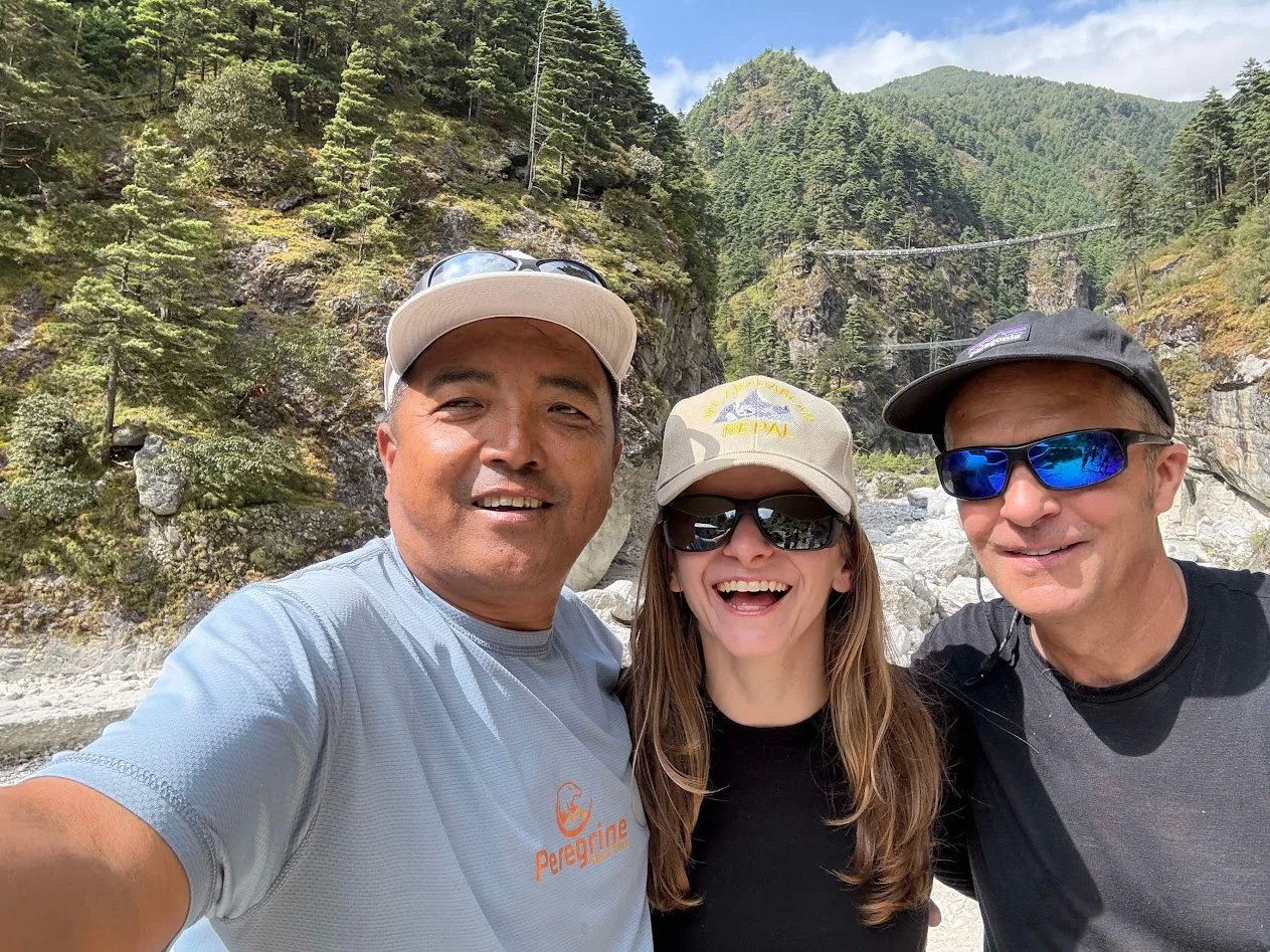 Three happy trekkers posing for a selfie during the Everest Base Camp trek fitness level assessment in the Khumbu valley, featuring a high suspension bridge and lush pine forests in the background.