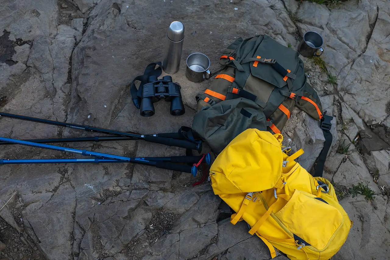 Essential Everest Base Camp trekking gear including backpack, trekking poles, binoculars, thermos, and camping mugs placed on rocky ground.