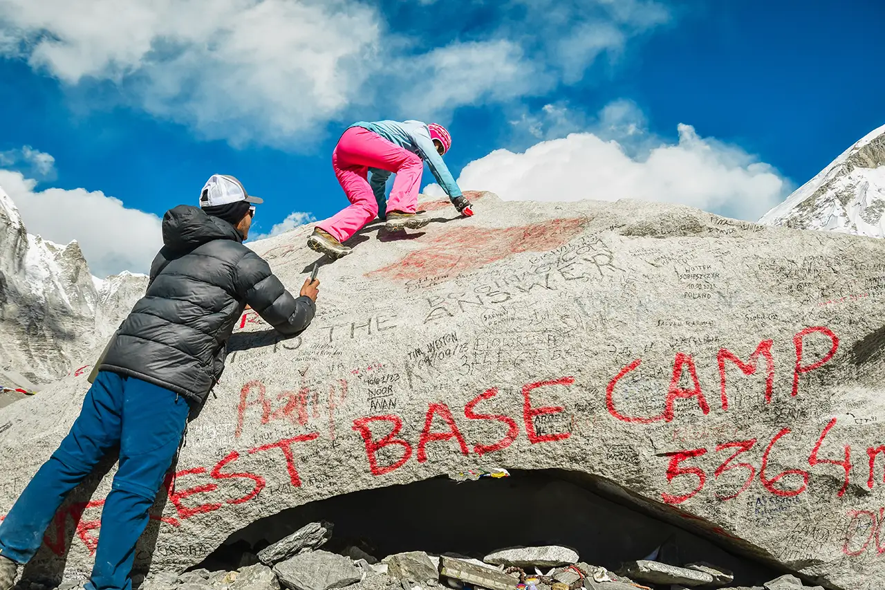A female trekker in pink pants climbing the iconic Everest Base Camp rock marked "Everest Base Camp 5364m" in red paint, while a guide takes a photo under a bright blue sky.