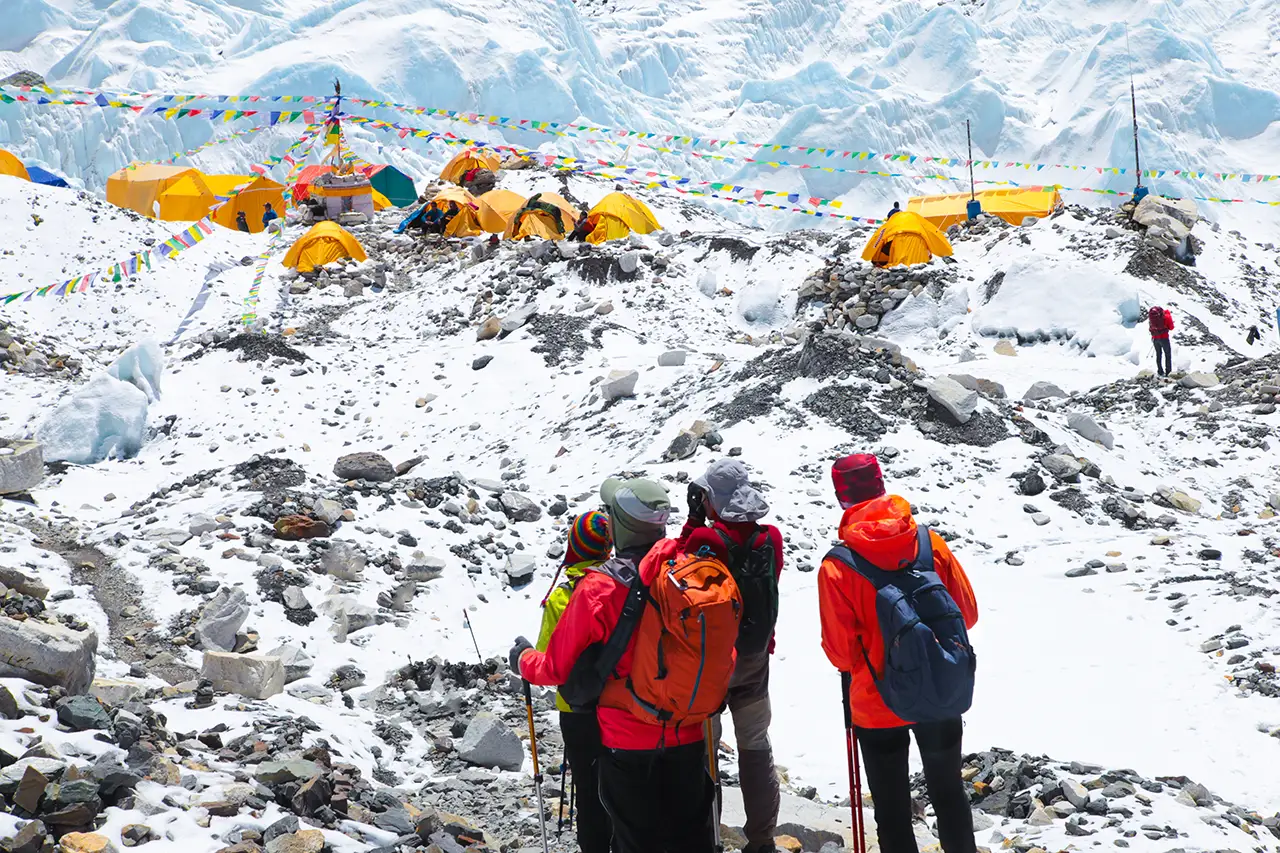 A group of four trekkers in bright red and orange jackets looking toward the yellow climber tents and prayer flags at Everest Base Camp, with the Khumbu Icefall in the background.