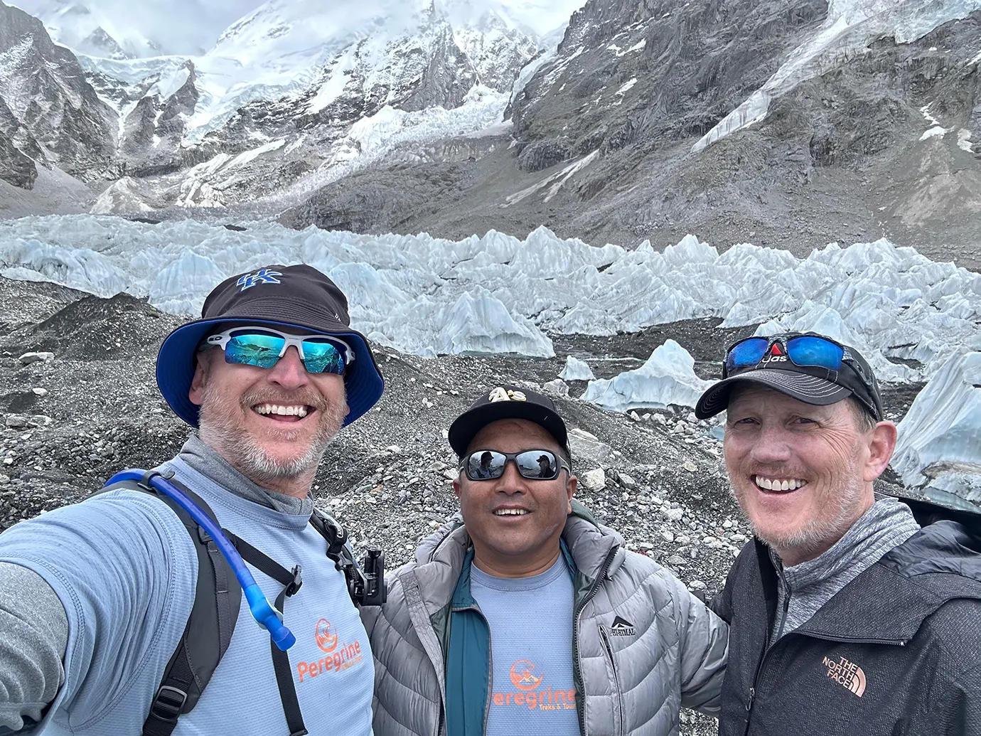 A smiling selfie of two hikers and their private guide to the Everest Base Camp Trek in front of the spectacular ice seracs of the Khumbu Glacier.