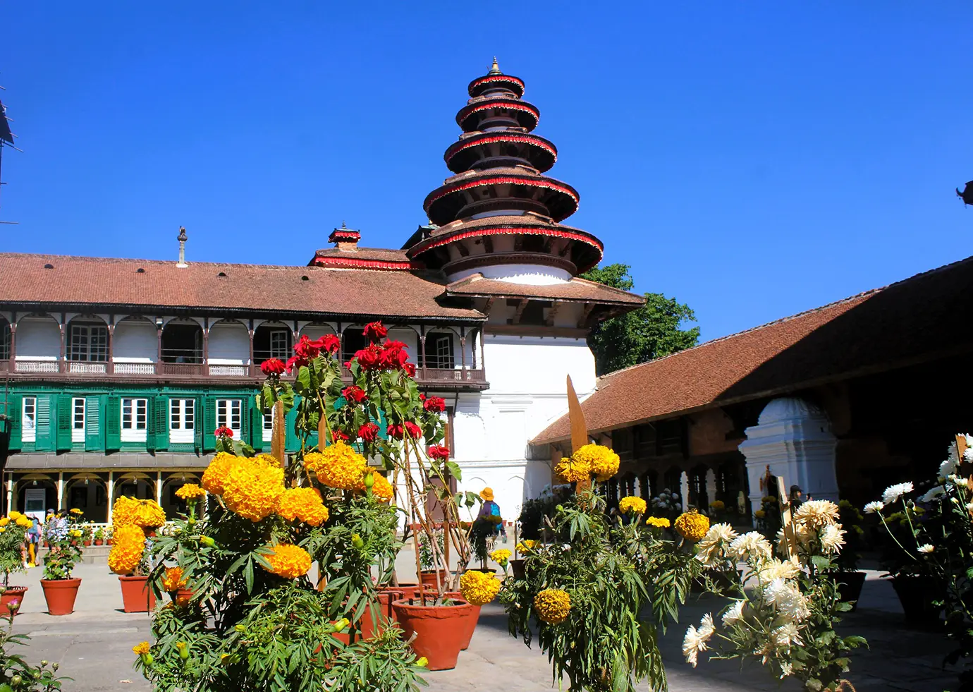 A high-resolution photo from Nasal Chowk, completely covered by a dense sea of potted marigolds, roses, and chrysanthemums forming a maze. In the background, the unique multi-tiered Chyasin Dega temple, the green-shuttered white palace building, and a legible sign mark the area as the 'Nasal Chowk Floral Maze'.