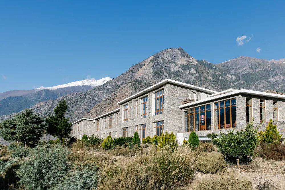Shinta Mani Mustang lodge exterior with stone architecture and large windows set against rugged Himalayan mountains and clear blue sky.