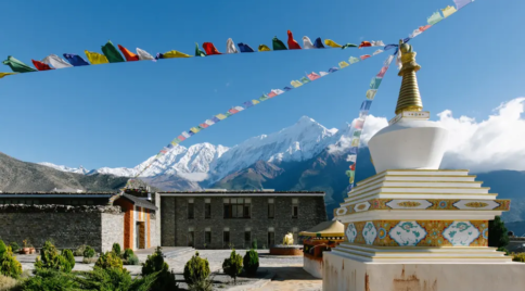 White stupa with prayer flags near a lodge building with snow-capped mountains in the background.
