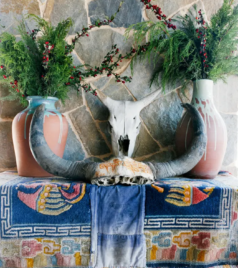 Traditional decorative skull with horns placed on a table with vases and greenery against a stone wall.