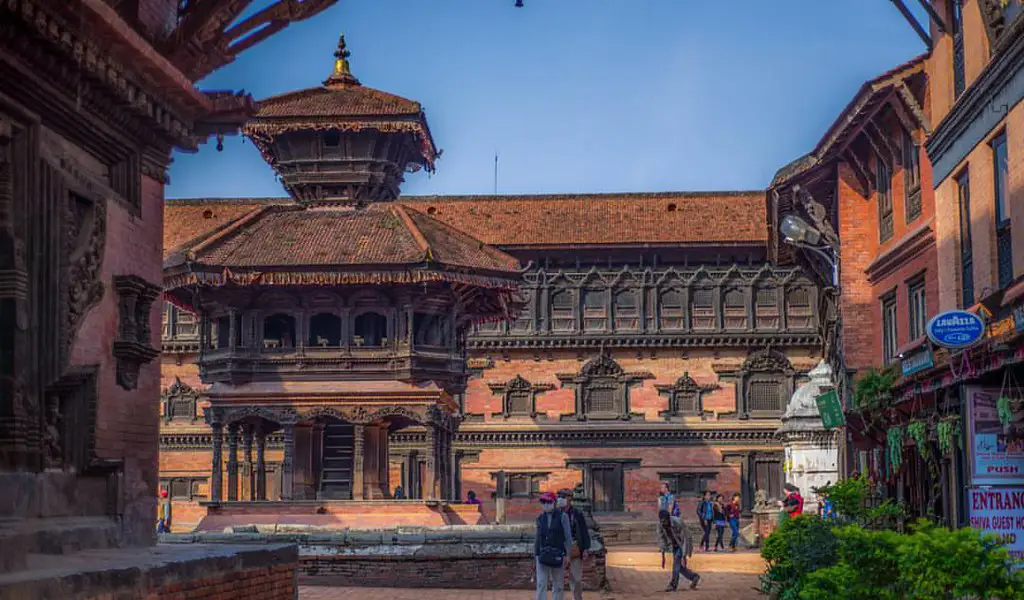 A view of traditional multi-tiered Newari temples and a palace building within a historic brick square in Kathmandu, Nepal. Tourists walk among ancient architecture, part of our Luxury Kathmandu Pokhara Chitwan Tour.