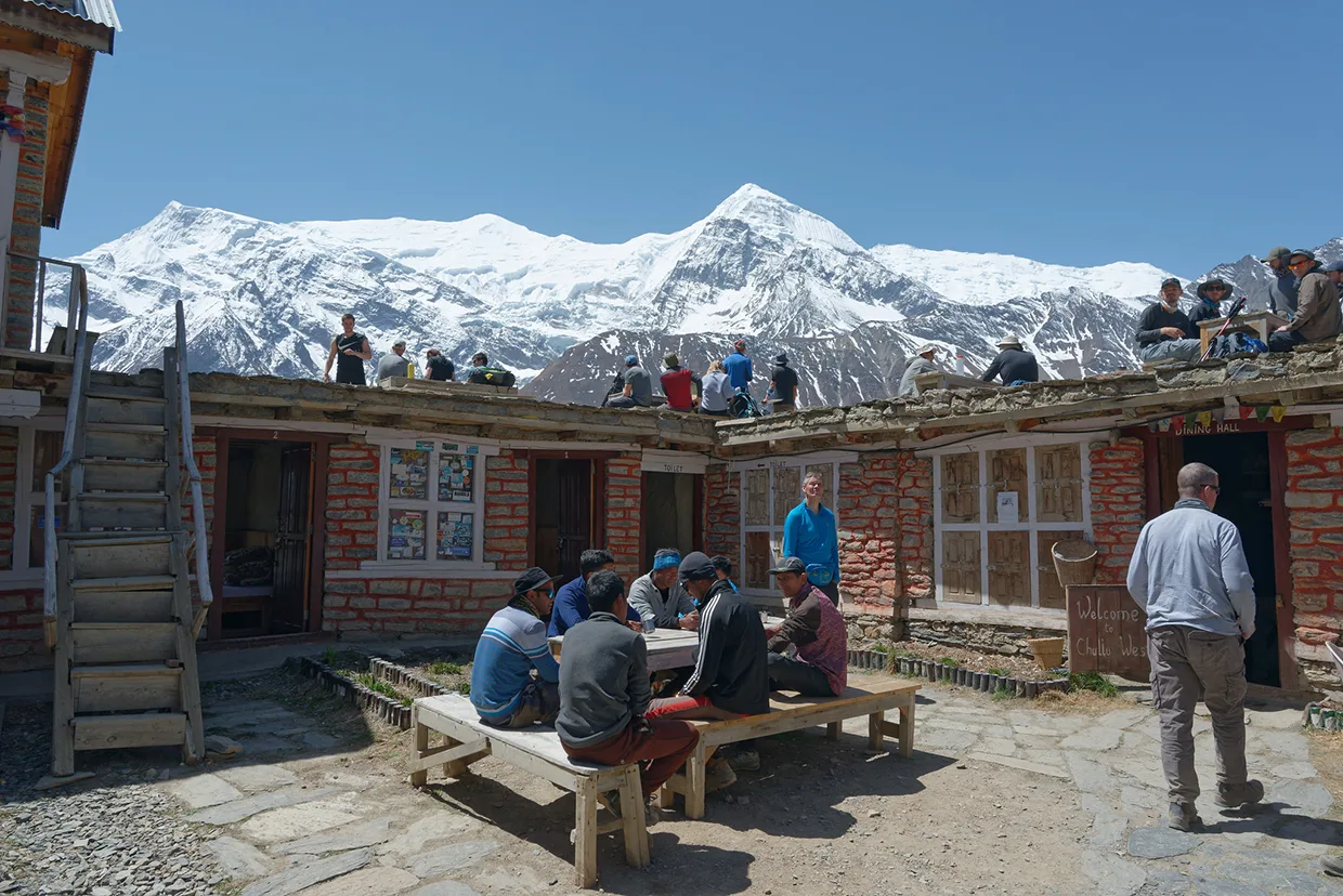 Trekkers relax outside a stone mountain teahouse in Yak Kharka with a stunning backdrop of snow-capped peaks during a Luxury Annapurna Circuit Trek.