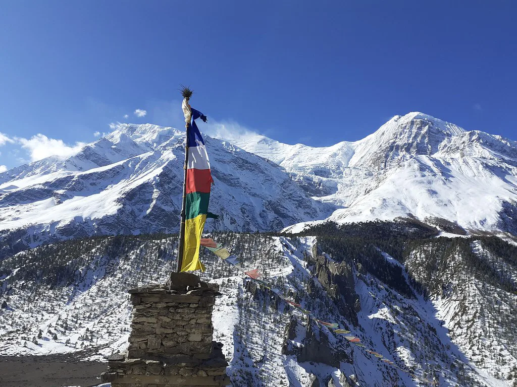 Colorful Buddhist prayer flags flutter in the wind against a backdrop of massive snow-covered Himalayan peaks on a Luxury Annapurna Circuit Trek.