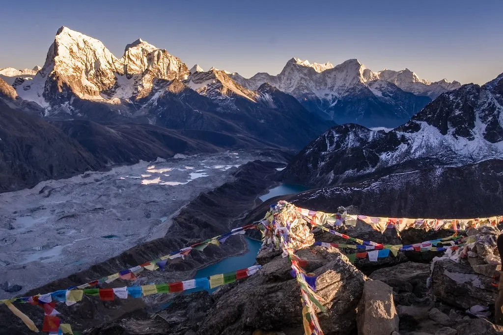 Colorful prayer flags flutter on a rocky summit overlooking the Ngozumpa Glacier and turquoise waters during a Luxury Everest Base Camp & Gokyo Lakes Trek.