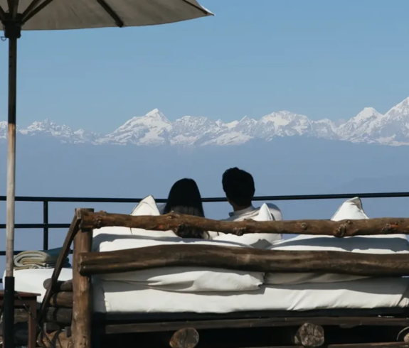 A couple relaxes on a plush outdoor daybed under a white umbrella, admiring the snow-capped Himalayan peaks during their Luxury Nepal Honeymoon Tour.
