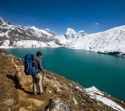 A solo hiker with a blue backpack overlooks the stunning turquoise waters of Gokyo Lake surrounded by snow-covered peaks during a Luxury Everest Base Camp & Gokyo Lakes Trek.