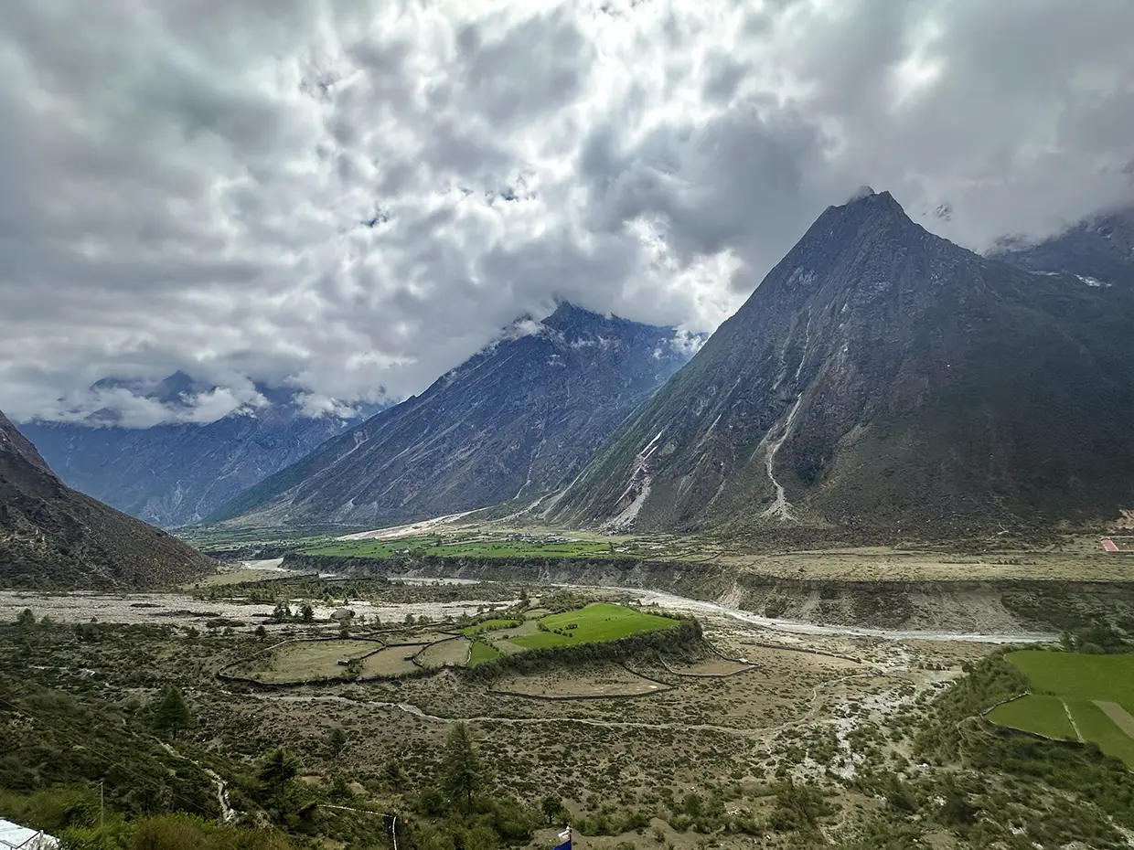 A panoramic view shows a wide glacial river valley with green agricultural fields surrounded by massive, steep mountain peaks and dramatic clouds during a Luxury Manaslu Trek.